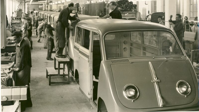Factory workers assembling vans on an auto production line in a black and white photo, reminiscent of "The Audi That Saved Audi: 60 Years Since Ingolstadt’s Turning Point," with partially completed vehicles lined up indoors.