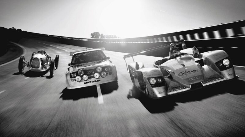 Three race cars from different eras drive side by side on a racetrack in black and white, with sunlight streaming in—a scene reminiscent of "The Audi That Saved Audi: 60 Years Since Ingolstadt’s Turning Point.