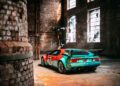 A vintage BMW sports car with a red and teal livery and number 76 is parked inside an old, sunlit brick warehouse with large windows—evoking the spirit of Monterey Car Week 2025.