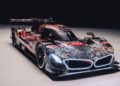 A modern 2025 BMW endurance race car with graphic livery, low body, wide tires, large rear wing, and illuminated front accents, pictured on a plain white floor in a dark studio—perfect for Monterey Car Week.