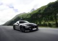 A BRABUS AMG GT speeds along a scenic mountain road, framed by green hills, trees, and a cloudy sky in the background.