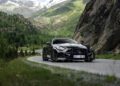 A black BRABUS AMG GT hybrid supercar drives on a winding mountain road, surrounded by greenery and rocky cliffs, with snow-capped peaks in the background.