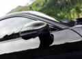 Close-up of a black AMG GT side mirror with a carbon fiber finish, reflecting the surroundings; blurred green trees and hills are visible in the background.