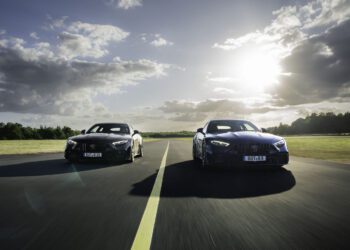 Two black sports cars drive side by side on an empty road under a partly cloudy sky with the sun shining in the background.