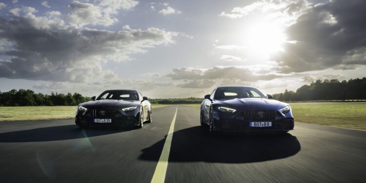 Two black sports cars drive side by side on an empty road under a partly cloudy sky with the sun shining in the background.