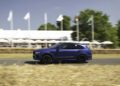 A blue Bentley Bentayga Speed drives on a road lined with straw bales at the Goodwood Hillclimb, with white tents, green trees, and several flagpoles in the background on a sunny day.