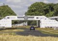 A blue Bentley Bentayga Speed drives on the Goodwood Hillclimb track at the Festival of Speed, passing under a white event arch with hay bales lining both sides and spectators in the background.