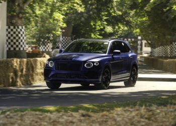 A blue Bentley Bentayga Speed luxury SUV drives on a paved road lined with hay bales and checkered barriers, surrounded by green trees in daylight, reminiscent of the famous Goodwood Hillclimb record runs.