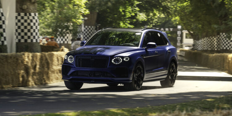A blue Bentley Bentayga Speed luxury SUV drives on a paved road lined with hay bales and checkered barriers, surrounded by green trees in daylight, reminiscent of the famous Goodwood Hillclimb record runs.