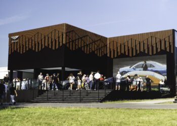 A group of people stand outside a modern Bentley Motors showroom at Goodwood, with a large screen displaying cars and a suited man, on a sunny day.