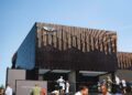 A modern black Bentley pavilion with geometric designs on the upper facade hosts a stylish car display by Bentley Motors, as people gather outside under a clear blue sky at Goodwood.