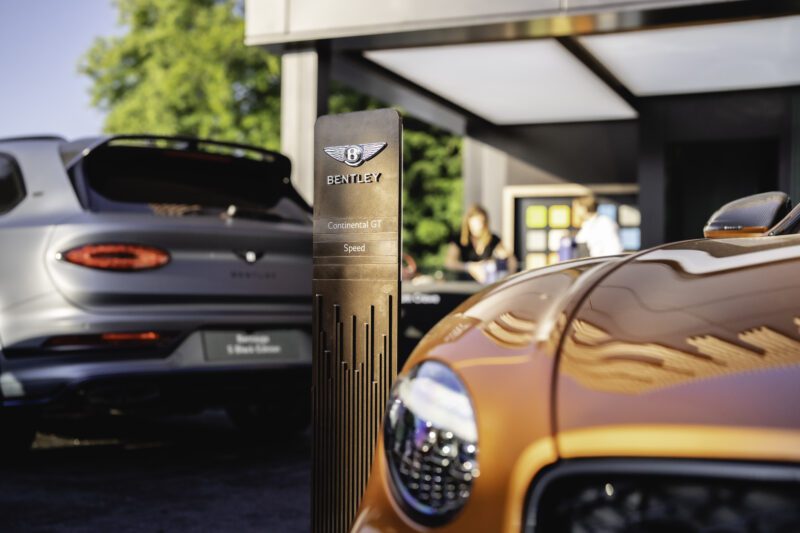 A close-up of an orange Bentley car at a Goodwood car display, next to a sign reading "Bentley Continental GT Speed," with another Bentley and blurred people in the background.