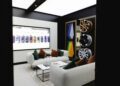 A modern showroom lounge with two white sofas, a coffee table, a Bentley Motors car display featuring wheels and paint samples, and a large screen showing an aerial view of parked cars.