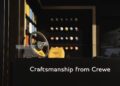 Steering wheel and orange headrest displayed in front of a wall with colorful car paint samples, with the text "Craftsmanship from Crewe" by Bentley Motors, featuring the largest display of finishes outside Goodwood.