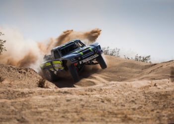 Off-road racing truck from Brenthel Industries kicks up dust while driving over a sandy dirt mound in a desert landscape, capturing the thrill of an Off-Road Adventure under clear skies.