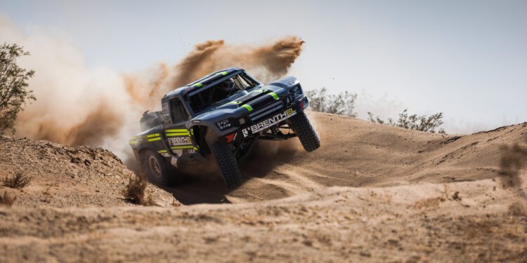 Off-road racing truck from Brenthel Industries kicks up dust while driving over a sandy dirt mound in a desert landscape, capturing the thrill of an Off-Road Adventure under clear skies.