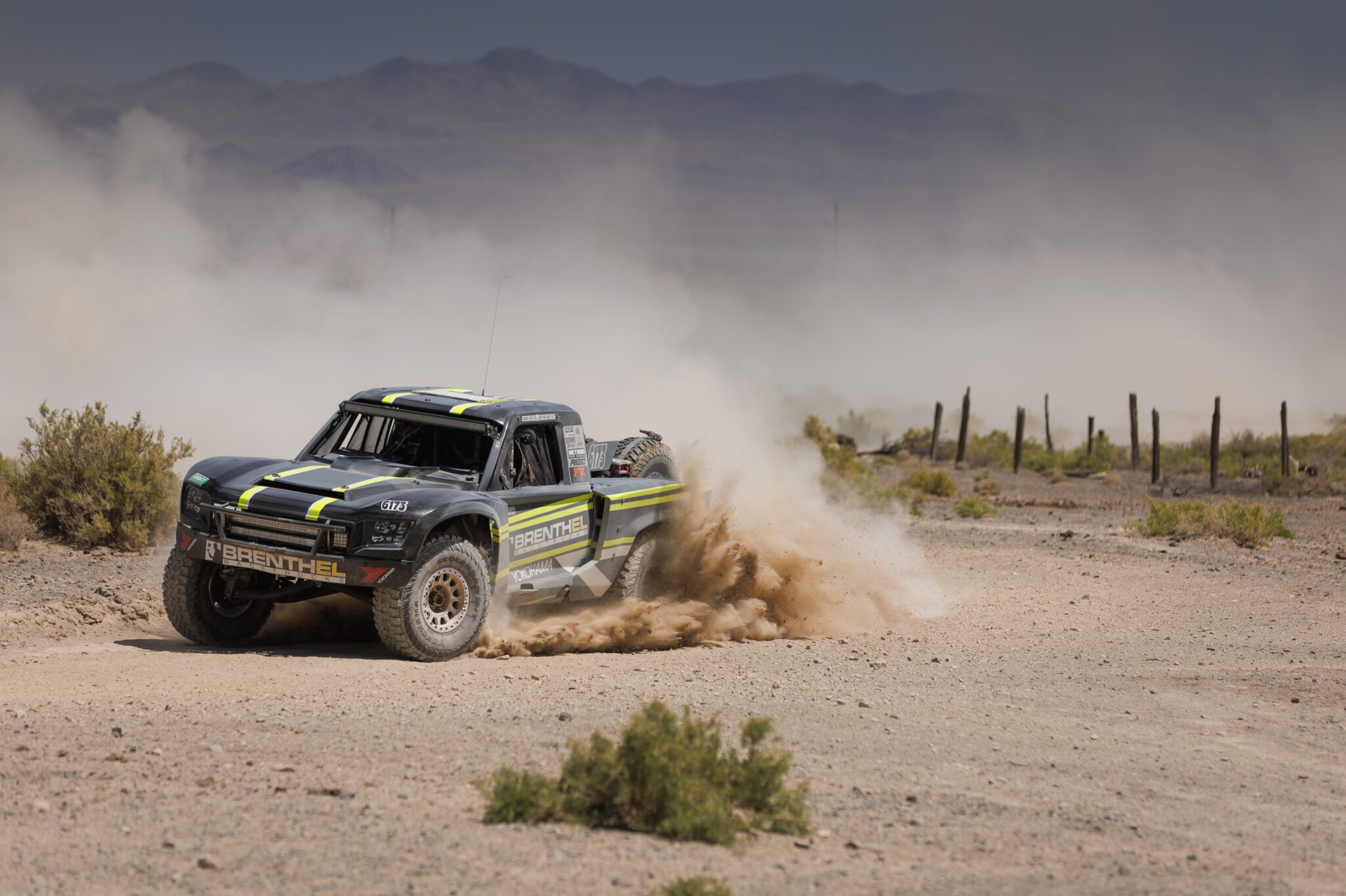 An off-road race truck from Brenthel Industries speeds through a dusty desert track, kicking up dirt, with mountains and sparse vegetation in the background.