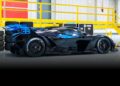 A black and blue race car with a large rear wing, believed to be the #001 of 40 Track-Only Bugatti Bolide set for auction debut at Pebble Beach, is parked indoors next to yellow railings and stacked tires.
