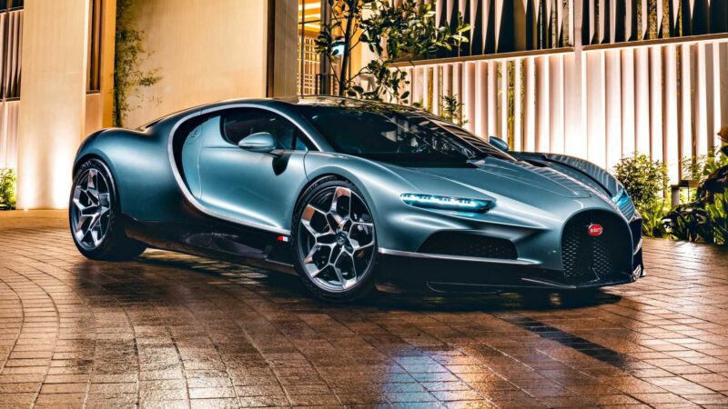 A silver Bugatti Chiron, one of the most powerful production cars, is parked on a tiled driveway at night, with modern building lights illuminating the background.