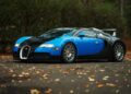 A blue and black Bugatti Veyron, one of the most expensive Bugatti hypercars of all time, is parked on a leaf-covered pavement with trees in the background.