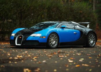 A blue and black Bugatti Veyron, one of the most expensive Bugatti hypercars of all time, is parked on a leaf-covered pavement with trees in the background.