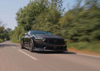 A Ford Mustang Dark Horse sports car speeds along a tree-lined, two-lane road under a clear sky, embodying the power and style of Clive Sutton’s renowned vehicles.