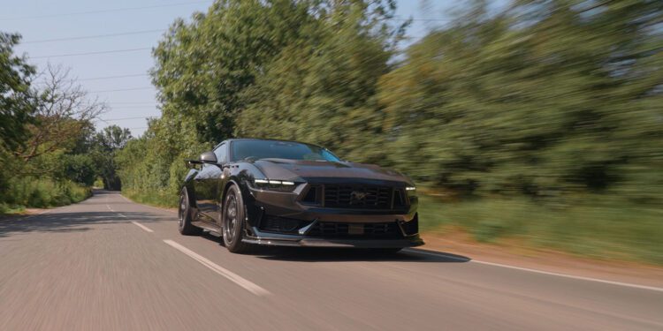 A Ford Mustang Dark Horse sports car speeds along a tree-lined, two-lane road under a clear sky, embodying the power and style of Clive Sutton’s renowned vehicles.