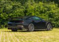 A black Ford Mustang Dark Horse with a rear wing is parked on a grassy area in front of dense green bushes.