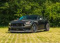 A black Ford Mustang Dark Horse sports car is parked on a grassy area with dense green trees in the background.