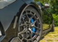 Close-up view of a Clive Sutton Ford Mustang Dark Horse sports car parked on a grassy, stone-covered surface, featuring blue brake calipers and black alloy wheels.