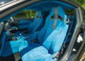 Interior view of a Ford Mustang featuring bright blue suede racing seats with black accents, center console, and door panels, photographed in daylight with greenery visible outside.