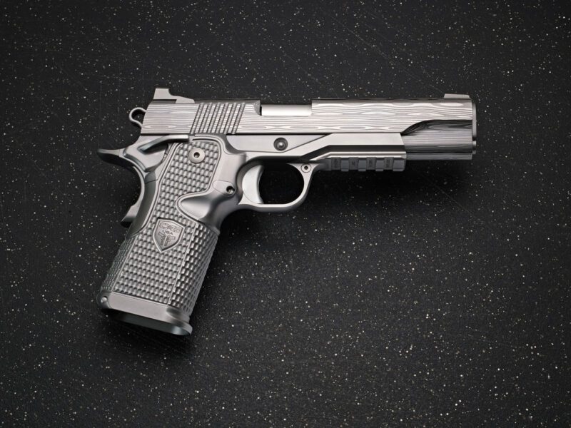 A silver Cabot Guns semi-automatic pistol with a textured grip rests on a black speckled surface, exuding both precision and legacy.