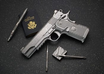 A silver-patterned Cabot Guns pistol, a U.S. passport, a pen, a metal lighter, and a metal tool arranged on a black speckled surface evoke an aura of legacy and precision.