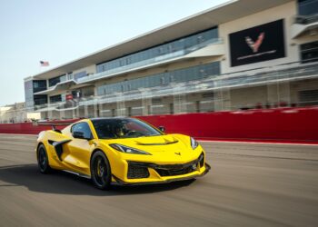 A yellow Track Review Corvette ZR1, proving 1000 HP doesn’t need a seven-figure price, speeds along a racetrack in front of a modern, multi-story building with large windows and a red barrier.