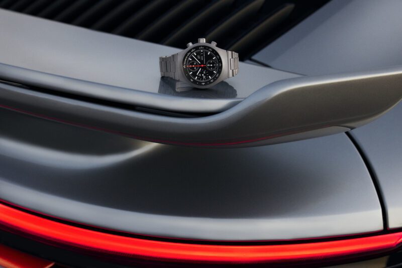 A Limited Edition wristwatch with a black dial and metal band is displayed on the rear spoiler of a silver sports car with a red taillight, showcasing classic Porsche Design elegance.