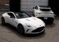 Two white Aston Martin cars, representing Cleveland Motorsports’ exquisite selection of exotic cars, are parked on cracked pavement in front of a closed garage door—one seen from the front, the other from the rear.