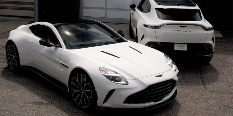 Two white Aston Martin cars, representing Cleveland Motorsports’ exquisite selection of exotic cars, are parked on cracked pavement in front of a closed garage door—one seen from the front, the other from the rear.