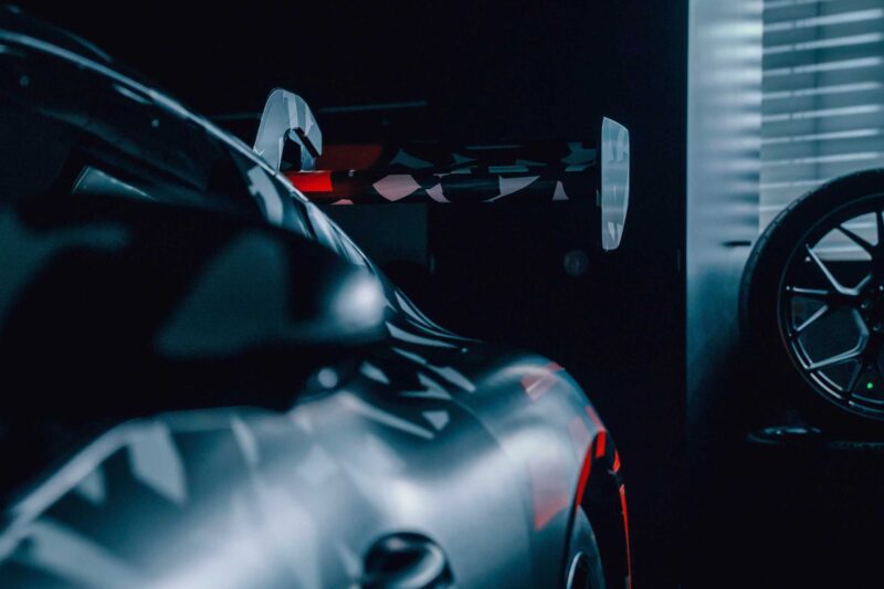 Close-up of the rear end of a Mercedes AMG GT with a large rear wing, partially in shadow, next to a window with blinds and a spare wheel—showcasing true track sport character.