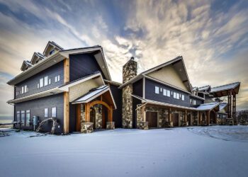 A large, modern house with stone accents and wooden beams stands in a snowy Alaska landscape under a partly cloudy sky, showcasing the golden potential of contemporary mountain living.