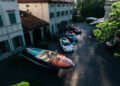 Aerial view of a courtyard with a wooden speedboat and several sports cars, including Ferraris from the Cavallino Classic, parked in front of a large house with green shutters.