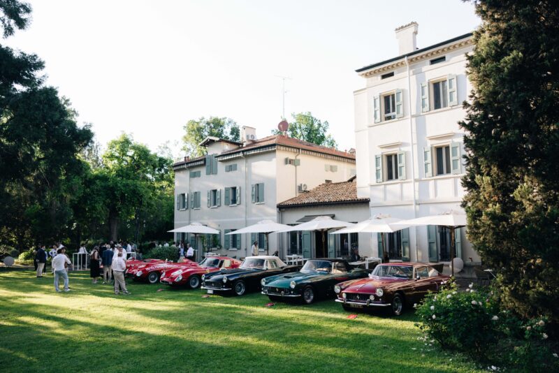 A row of classic cars is parked on a lawn outside a large white villa, with people standing nearby under umbrellas on a sunny day at the Premier Ferrari Event.