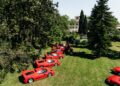 A row of red sports cars is parked on a grassy lawn at the Ferrari Concours, with people standing nearby and trees and a large building in the background.