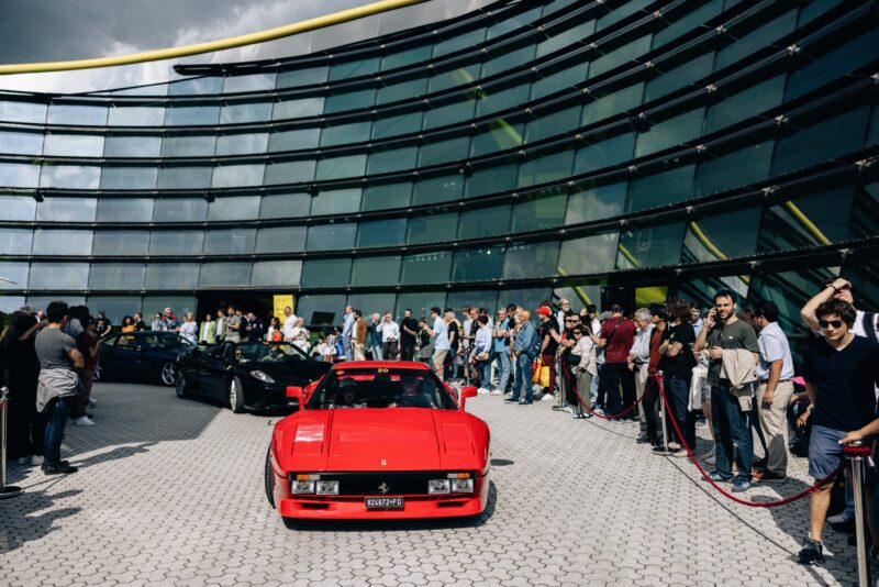 A red Ferrari drives past a crowd of people gathered outside a modern glass building on a cloudy day during the premier Ferrari event.