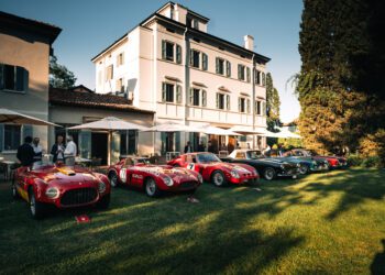 A row of classic sports cars is displayed on a lawn in front of a large, elegant white building at the renowned Cavallino Classic, with a few people standing nearby.