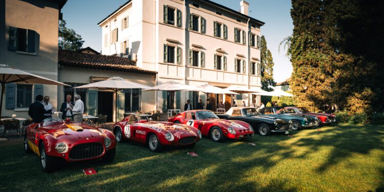 A row of classic sports cars is displayed on a lawn in front of a large, elegant white building at the renowned Cavallino Classic, with a few people standing nearby.