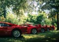 Four red sports cars are parked in a row on grass under trees, sunlight filtering through the leaves, capturing the elegance of a Ferrari Concours.