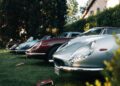 A row of classic sports cars, including a stunning Ferrari, is parked on grass beside a hedge, with sunlight filtering through the trees at this prestigious Concours Event.
