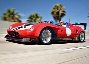 A red vintage-style Ferrari 612TR sports car with a white circle and black stripes is driven on a highway by a man wearing sunglasses; palm trees and blue sky in the background, capturing true V12 exotic flair.