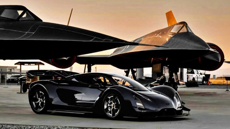 A sleek black 2025 sports car, one of the most powerful production cars, is parked in front of two black jet aircraft on an airfield at sunset.