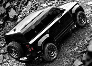 A 2026 Land Rover Defender in OCTA Black with a spare tire mounted on the rear drives up a rocky, uneven slope.